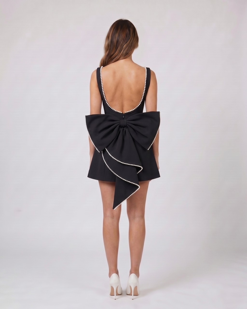 Back view of black wedding guest pearl trim back bow mini dress showing Deep U-shaped open back with pearl trim along edges, center back invisible zipper closure, and a large sculptural multi-layered bow with pearl-trimmed edges attached at the waist.