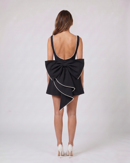 Back view of black wedding guest pearl trim back bow mini dress showing Deep U-shaped open back with pearl trim along edges, center back invisible zipper closure, and a large sculptural multi-layered bow with pearl-trimmed edges attached at the waist.