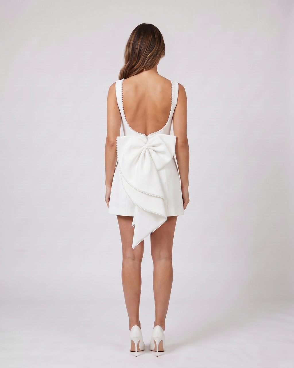 Back view of white wedding guest pearl trim back bow mini dress showing Deep U-shaped open back with pearl trim along edges, center back invisible zipper closure, and a large sculptural multi-layered bow with pearl-trimmed edges attached at the waist.