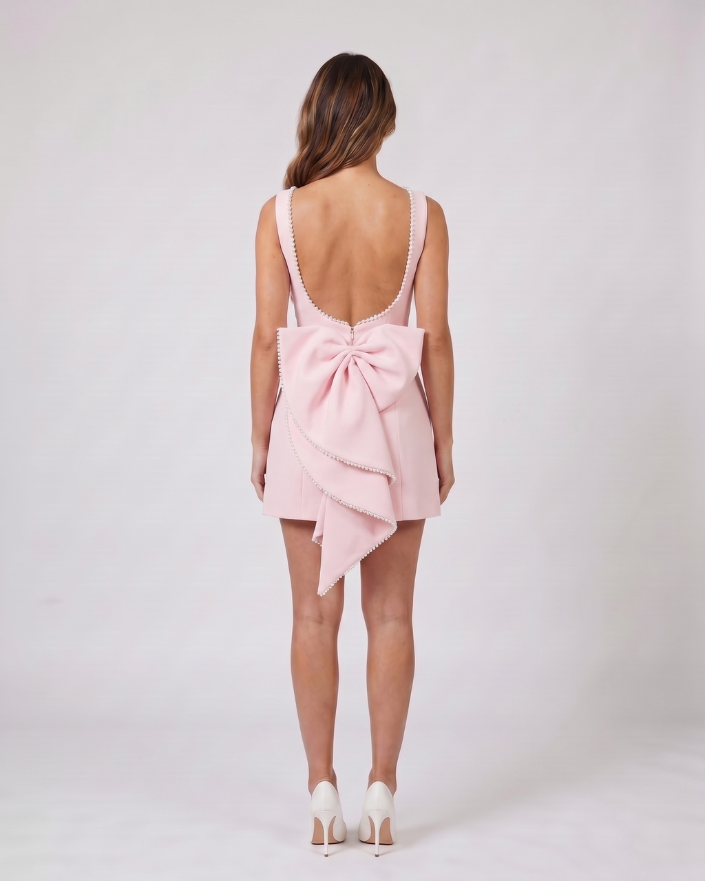 Back view of pink wedding guest pearl trim back bow mini dress showing Deep U-shaped open back with pearl trim along edges, center back invisible zipper closure, and a large sculptural multi-layered bow with pearl-trimmed edges attached at the waist.